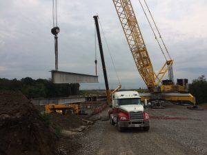 A crane is seen in view dropping down a girder into place. A semi-truck can be seen in the foreground.